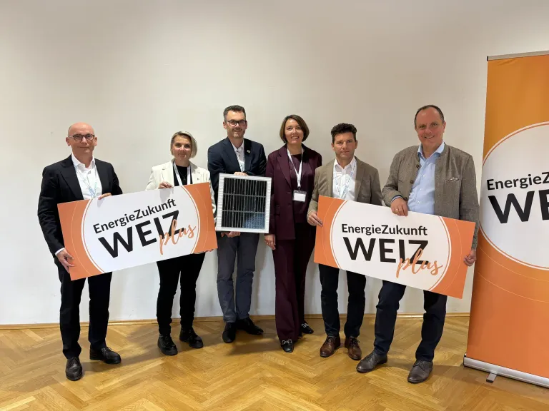 Fünf Personen halten Schilder mit 'EnergieZukunft WEIZ plus'. Sie lächeln für ein Foto. Dahinter ist eine weiße Wand und ein Holzboden.