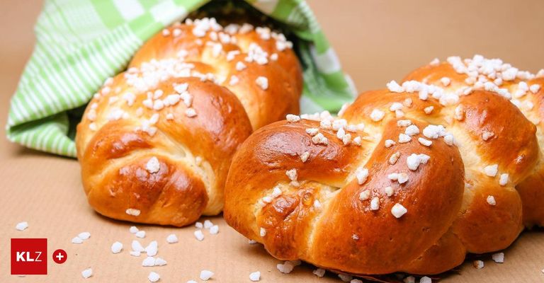 Two braided breads with white toppings are placed on a brown surface. They are sprinkled with white crystals. A green cloth is partially covering the bread.