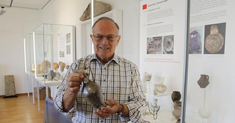 Ein älterer Mann lächelt und hält eine antike Vase in einem Museum, umgeben von anderen historischen Artefakten.