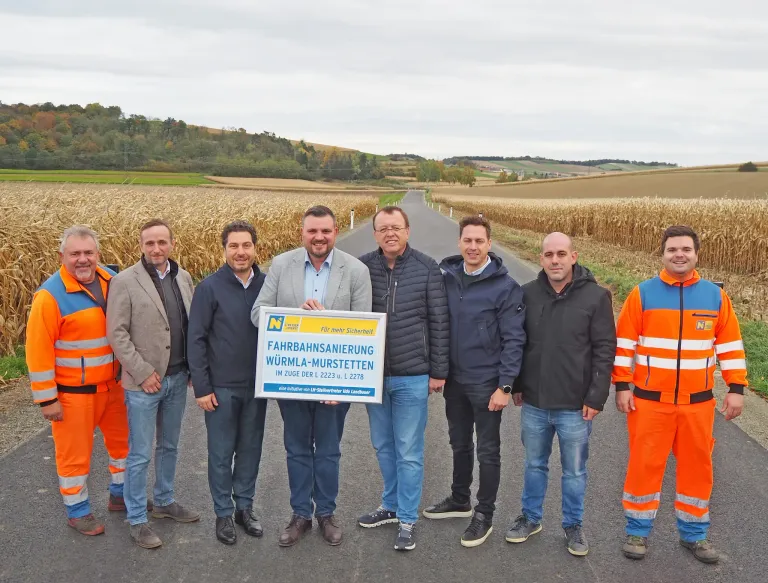Eine Gruppe von Männern, gekleidet in reflektierende Westen und formelle Kleidung, steht auf einer ländlichen Straße. Sie halten ein Schild mit der Aufschrift 'FAHRBAHNSANIERUNG WÜRMLÄ-MÜRSTETTEN'. Der Hintergrund zeigt weite Felder und einen bewölkten Himmel.