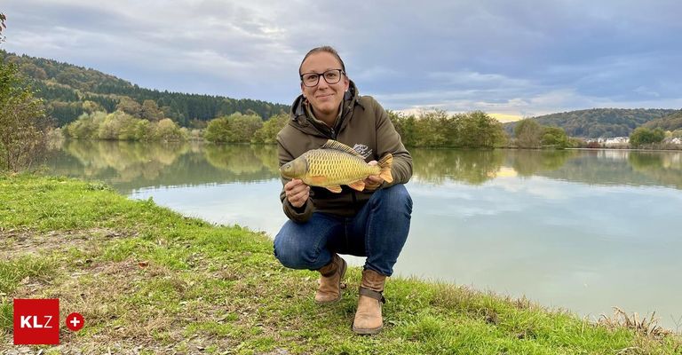 Eine Frau in einer braunen Jacke und mit Brille kniet am Ufer eines Sees und hält einen goldenen Fisch. Hinter ihr spiegelt der See den Himmel und die Bäume wider.