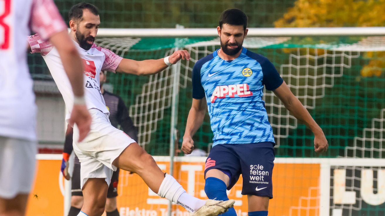 Zwei Fußballspieler führen ein dynamisches Spiel auf einem Feld, einer in Weiß und der andere in Blau. Sie stehen vor einem Netz, wobei ein dritter Spieler teilweise im Hintergrund zu sehen ist.