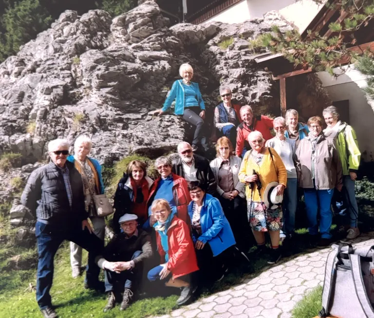 Eine Gruppe von Menschen posiert für ein Foto vor einer großen Felsformation, einige stehend und andere auf dem Gras sitzend. Eine Frau steht auf dem Felsen. Bäume und ein Gebäude sind im Hintergrund zu sehen.