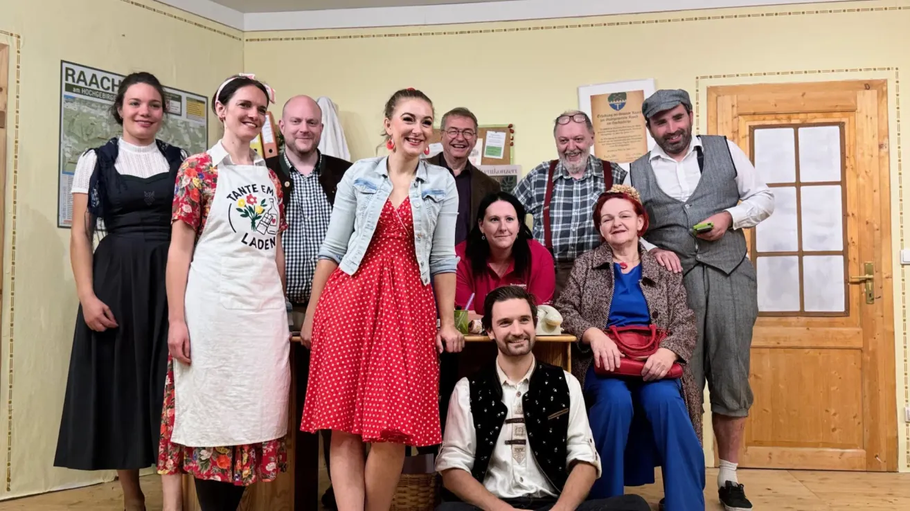 Eine Gruppe von Menschen in einem Innenraum posiert für ein Foto. Sie stehen und sitzen, einige lächeln. Eine Frau in einem roten Kleid mit Punkten steht vorne. Hinter ihr sitzt ein Mann in einem weißen Hemd. Auf der linken Seite trägt eine Frau ein Blumenkleid und eine Schürze.