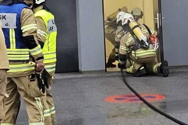 Feuerwehrleute in Schutzkleidung trainieren in der Nähe einer Tür, einer von ihnen trägt einen Feuerlöscher. Ein anderer kniet mit einer Feuerwehrspritze. Die Szene beinhaltet nasse Bodenmarkierungen.
