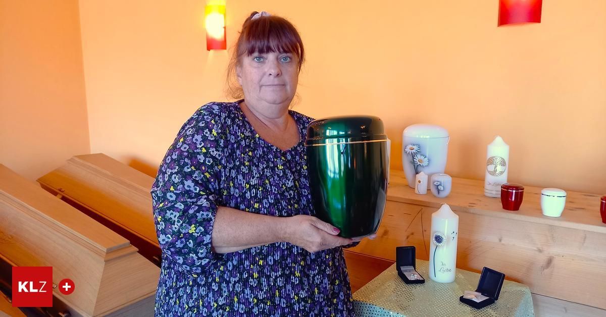 Eine Frau hält eine grüne Urne in einem Raum mit Holzregalen, die dekorative Gegenstände wie weiße Keramikvasen und Kerzen aufweisen, was auf eine Umgebung für Gedenk- oder Bestattungsarrangements hindeutet.