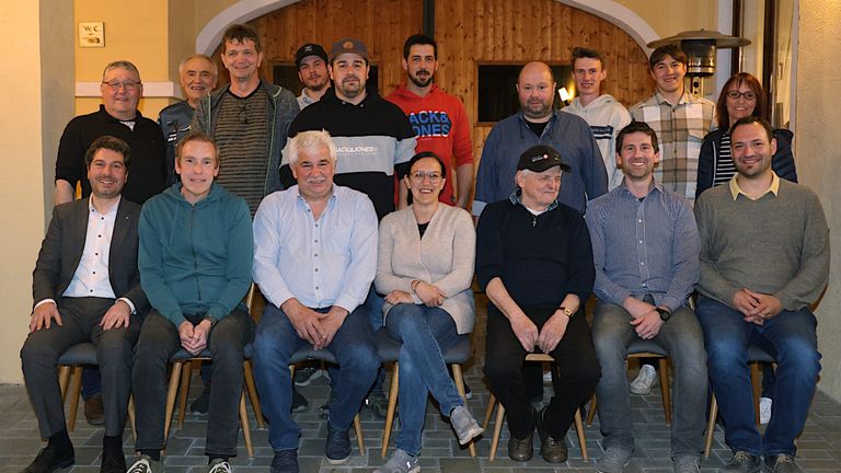 Eine Gruppe von Menschen posiert für ein Foto. Sie sitzen auf Stühlen und stehen dahinter. Eine Frau trägt eine Brille. Dahinter ist eine Holzwand.