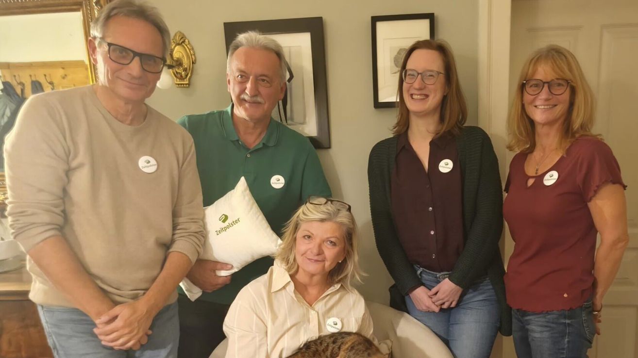 Vier Personen stehen eng beieinander drinnen. Ein Mann hält ein Kissen mit 'Zeitpolster' darauf. Eine andere Person hat eine Katze auf dem Schoß. An der Wand hinter ihnen hängen zwei gerahmte Bilder.