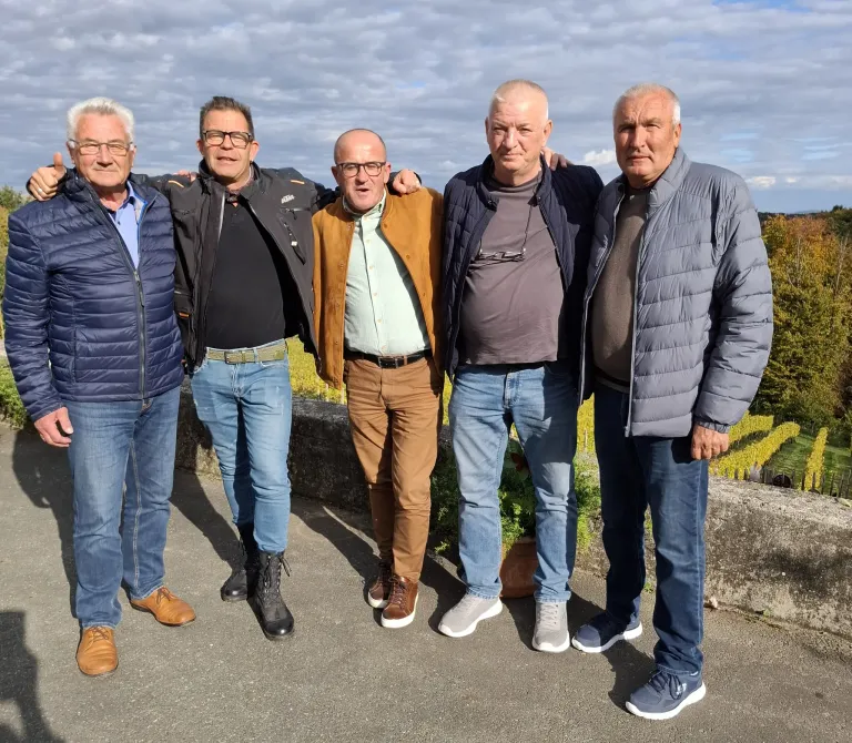 Fünf Männer stehen zusammen in einem Weinberg. Sie tragen alle Jacken und Jeans. Ein Mann trägt braune Hosen. Sie haben ihre Arme um die Schultern der anderen gelegt.