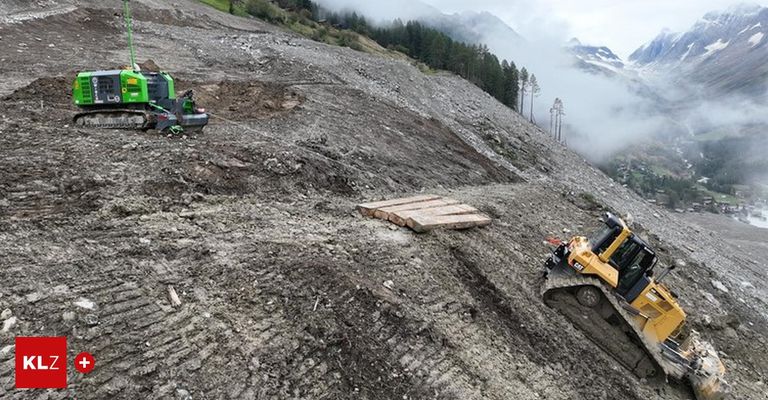 Eine Baustelle mit einem Baufahrzeug am Hang, umgeben von Kiefern und einem nebligen Berg im Hintergrund.
