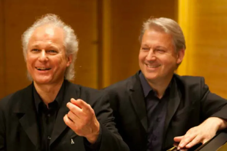 Zwei Männer mit weißen Haaren lächeln und schauen in die Kamera. Sie tragen schwarze Anzüge. Der Mann auf der linken Seite gestikuliert mit der Hand.
