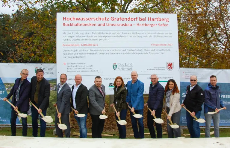 Eine Gruppe von Menschen steht vor einem Schild, das Hochwasserschutz Grafendorf bei Hartberg Rückhaltebecken und Linienausbau Hartberger Safen besagt. Sie halten Schaufeln und scheinen sich auf eine Veranstaltung vorzubereiten. Das Schild weist auf ein Hochwasserschutzprojekt hin, das von verschiedenen Organisationen unterstützt wird.