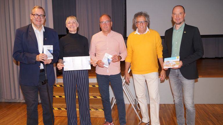 Fünf Erwachsene stehen in einer Reihe und halten Urkunden. Sie alle lächeln. Hinter ihnen befindet sich eine Treppe mit einer weißen Wand.