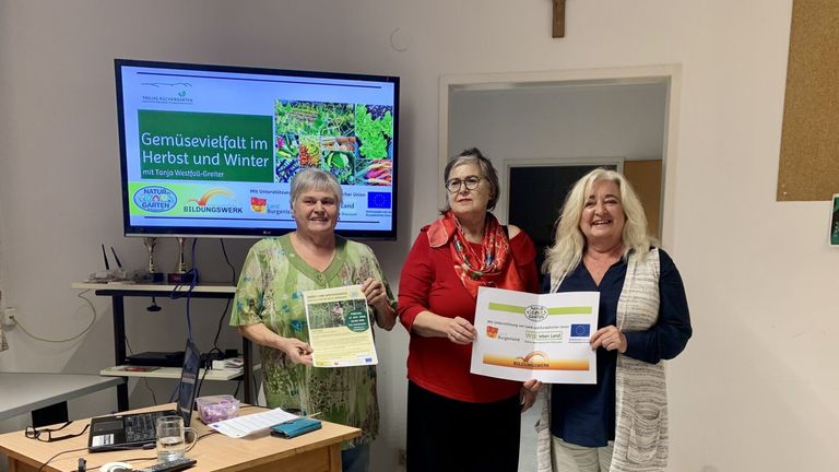 Drei Frauen stehen lächelnd in einem Raum und halten Urkunden. Hinter ihnen zeigt ein Monitor einen grünen Bildschirm mit Text über Vielfalt im Winter. Ein Kreuz ist an der Wand sichtbar.