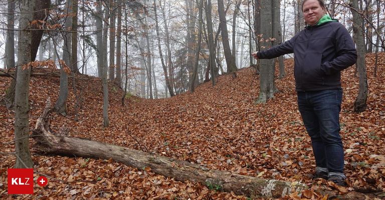 Ein Mann in einer Jacke steht in einem nebligen Wald und zeigt mit seiner rechten Hand. Der Boden ist mit gefallenen Blättern bedeckt, und die Bäume sind kahl.