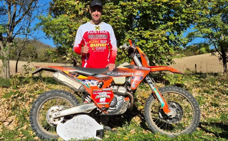 Ein Mann steht neben einem orangefarbenen Motorrad mit dem Wort Champion darauf, er trägt ein rot-weißes Shirt mit Logos.