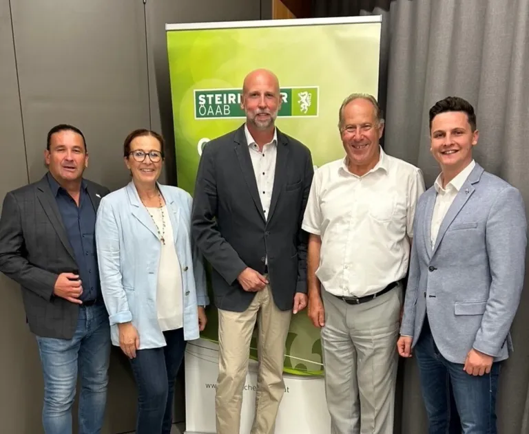 Fünf Personen stehen neben einem Banner mit den Worten 'Steirer Oaab'. Sie sind alle in formeller Kleidung gekleidet.