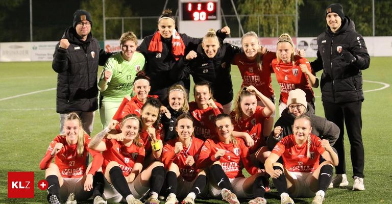 Eine Frauenfußballmannschaft in roten Trikots feiert auf einem Feld, wobei die Anzeigetafel 1:0 anzeigt, während einige Spielerinnen Brillen und Mützen tragen.