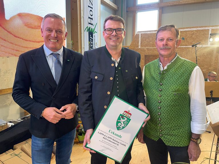 Drei Männer stehen zusammen in einem Raum. Der Mann in der Mitte hält ein gerahmtes Zertifikat. Der Mann auf der linken Seite trägt einen Anzug und eine Krawatte, während der Mann auf der rechten Seite eine grüne Weste trägt. Sie alle lächeln und scheinen für ein Foto zu posieren. Hinter ihnen ist eine Wand mit einer Holzstruktur und einer Pflanze.