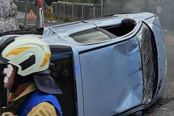 Ein Feuerwehrmann steht neben einem beschädigten Auto am Straßenrand, aus dem Rauch austritt. Ein Felsen und ein Metallzaun sind im Hintergrund zu sehen.
