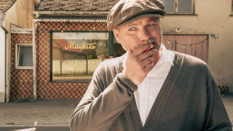 Ein Mann in Hut und Strickjacke hält einen roten Stift an seinen Lippen vor einem Geschäft mit Glasdisplay und dem Namen Maikits.