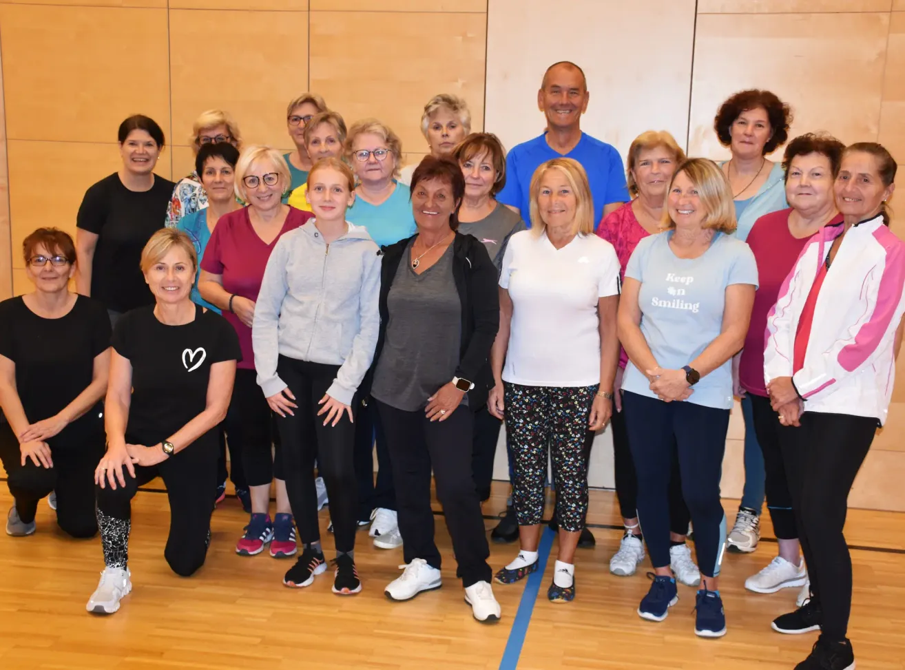 Eine Gruppe lächelnder Erwachsener in Trainingskleidung, stehend und kniend in einem Fitnessstudio. Eine Frau in der Mitte trägt eine Halskette. Eine Frau auf der rechten Seite trägt ein Shirt mit 'Keep on Smiling'.