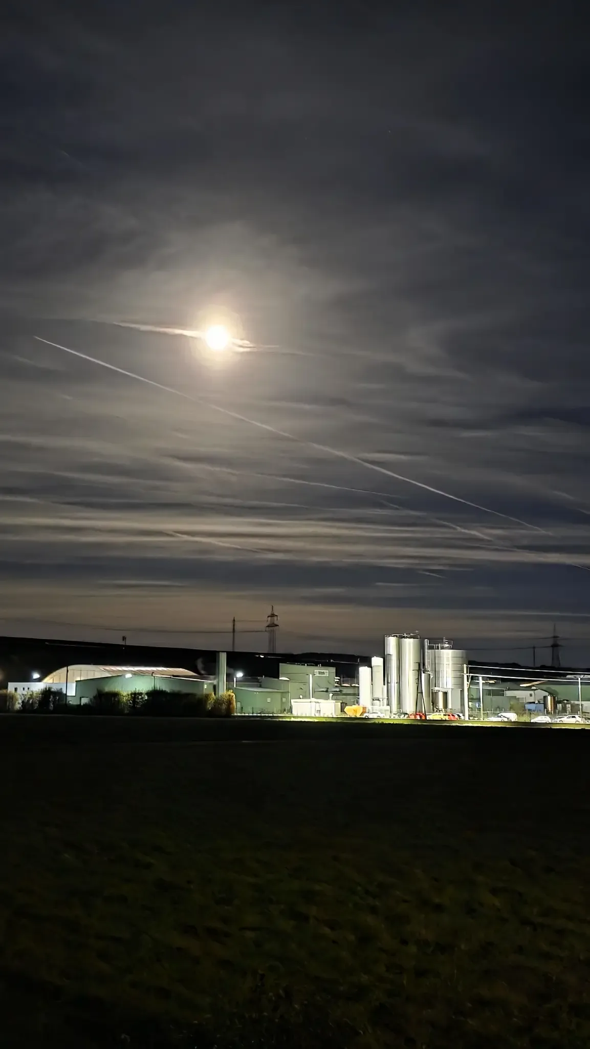 Eine mondbeschienene Nacht mit Industriegebäuden, großen Tanks und einem Turm im Hintergrund, mit langen Kondensstreifen, die den Himmel durchkreuzen.