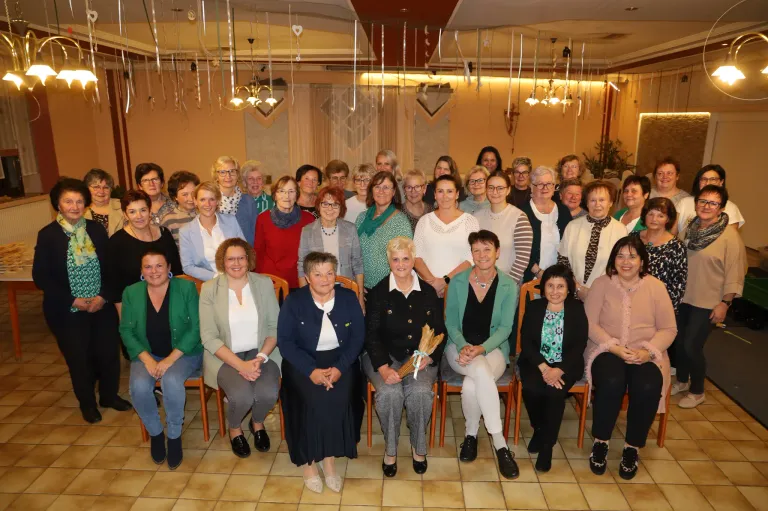 Eine Gruppe von Frauen, möglicherweise Kolleginnen oder Freundinnen, posiert für ein Foto in einem Raum mit gefliesten Böden, trägt verschiedene Outfits und einige halten Blumensträuße.