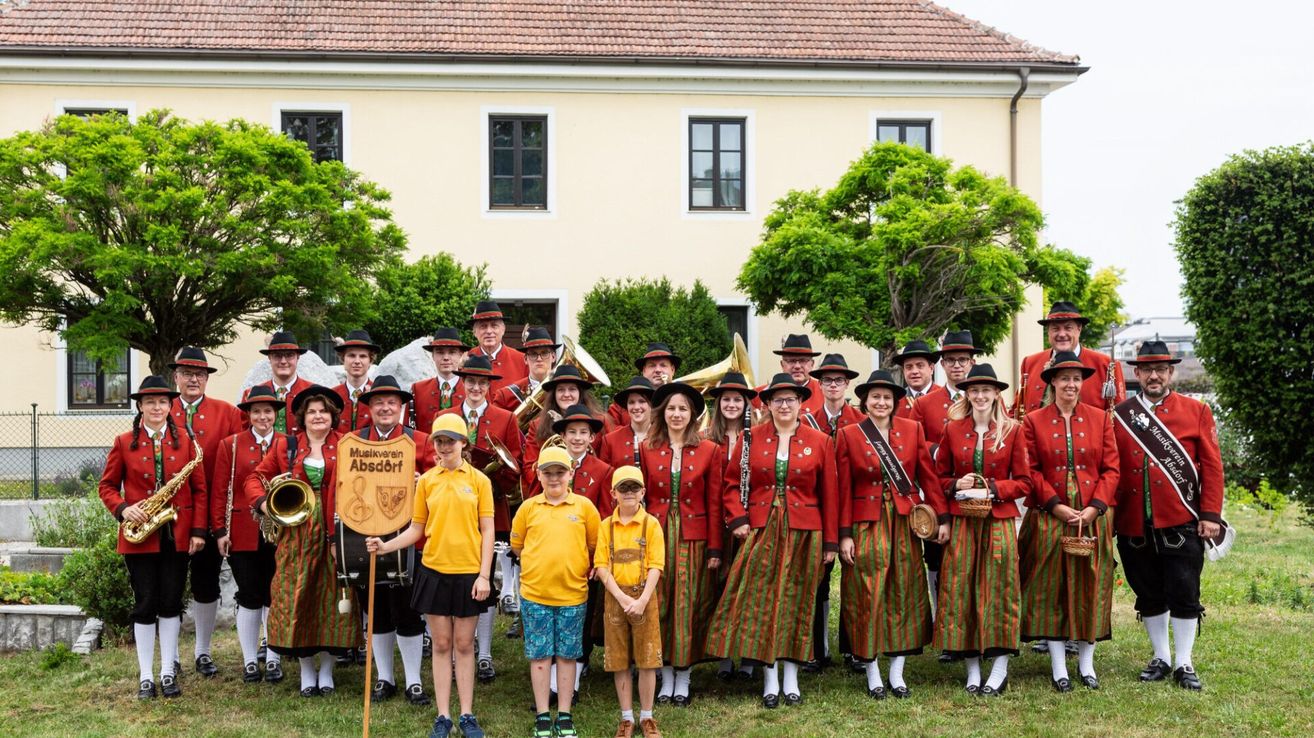 Eine Gruppe von Musikern, sowohl Erwachsene als auch Kinder, posiert vor einem Gebäude mit traditionellen Kostümen, Musikinstrumenten und lächelnden Gesichtern.