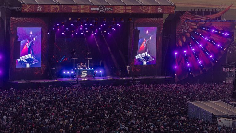 A live concert at night with a stage and an audience, featuring a guitarist on a screen and purple lights.