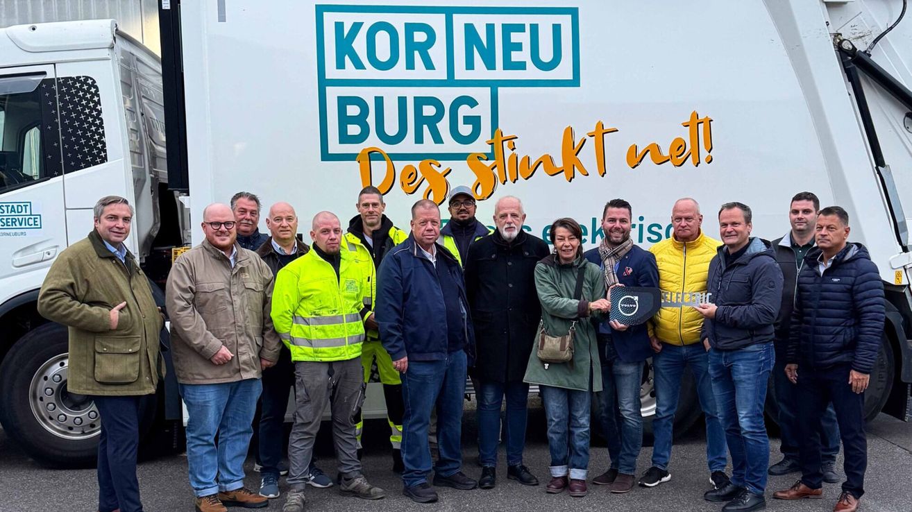 Eine Gruppe von Menschen in verschiedenen Jacken und Mützen steht vor einem Lastwagen mit einem Logo und Text darauf.