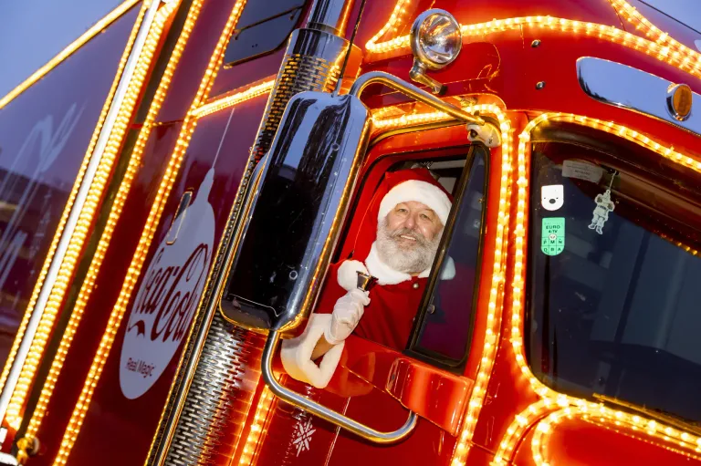 Ein Mann in Santa-Claus-Kleidung sitzt in einem roten Coca-Cola-Truck mit Weihnachtsbeleuchtung. Er hält eine Glocke und lächelt.