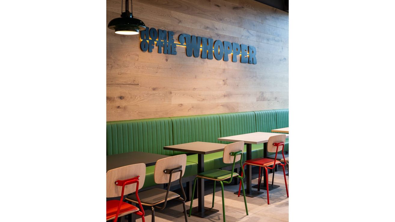 Innenansicht eines Whopper-Themenrestaurants mit Holzwänden und grünen Bänken. Tische und Stühle sind angeordnet. Das Schild 'HOME OF THE WHOPPER' ist beleuchtet.