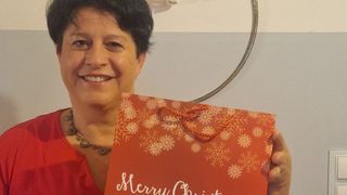 Eine Frau lächelt und hält einen roten Merry Christmas Geschenkbeutel mit weißen Schneeflocken. Sie trägt eine Halskette. Hinter ihr ist eine weiße Wand mit einem silbernen kreisförmigen Objekt.
