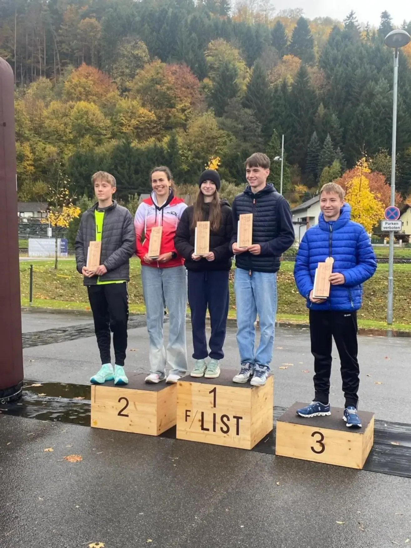 Fünf junge Menschen stehen auf Podesten und halten Holztafeln in den Händen. Sie befinden sich in einem Außenbereich mit Bäumen und Gras im Hintergrund. Die Podeste sind mit 1, 2 und 3 nummeriert.