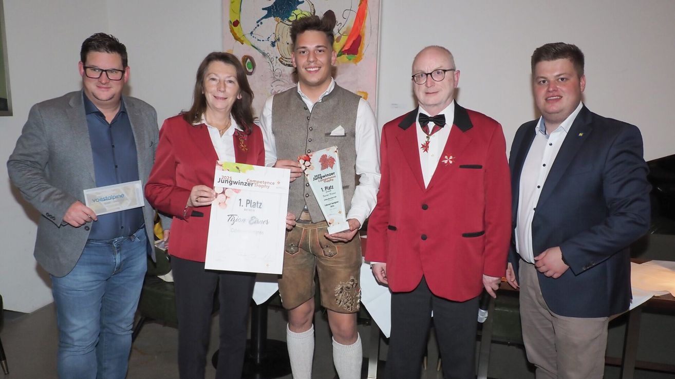 Drei Personen stehen nebeneinander und halten Urkunden. Ein Mann in traditioneller bayerischer Kleidung hält eine Urkunde für den 1. Platz. Ein Mann in einem roten Jackett und Fliege steht neben ihm. Eine Frau in einem roten Jackett hält eine Urkunde mit 'Jungwinzer-Kompetenz-Trophy' darauf.