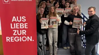 Eine Gruppe von Schülern und ein Mann posieren für ein Foto vor einem roten Banner, auf dem 'AUS LIEBE ZUR REGION' steht. Die Schüler halten Zeitungen in der Hand und lächeln. Der Mann macht eine Daumen hoch.
