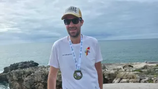 Ein Mann steht an einer Betonbarriere am Meer, trägt einen Hut, eine Sonnenbrille und eine Medaille um den Hals. Er lächelt und lehnt sich an die Barriere.