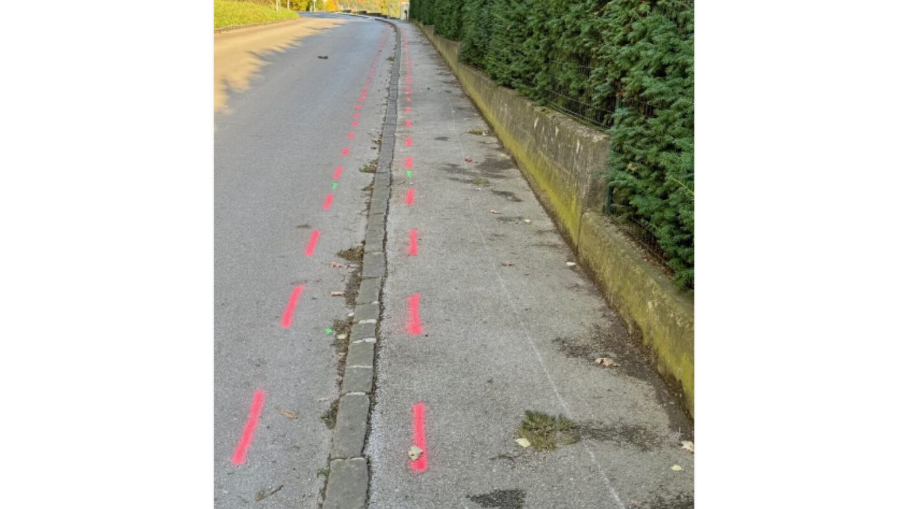 Eine Straße mit bemalten Linien auf dem Bürgersteig, die zu einem Steinrand führen, umgeben von Grün und einer Steinmauer.