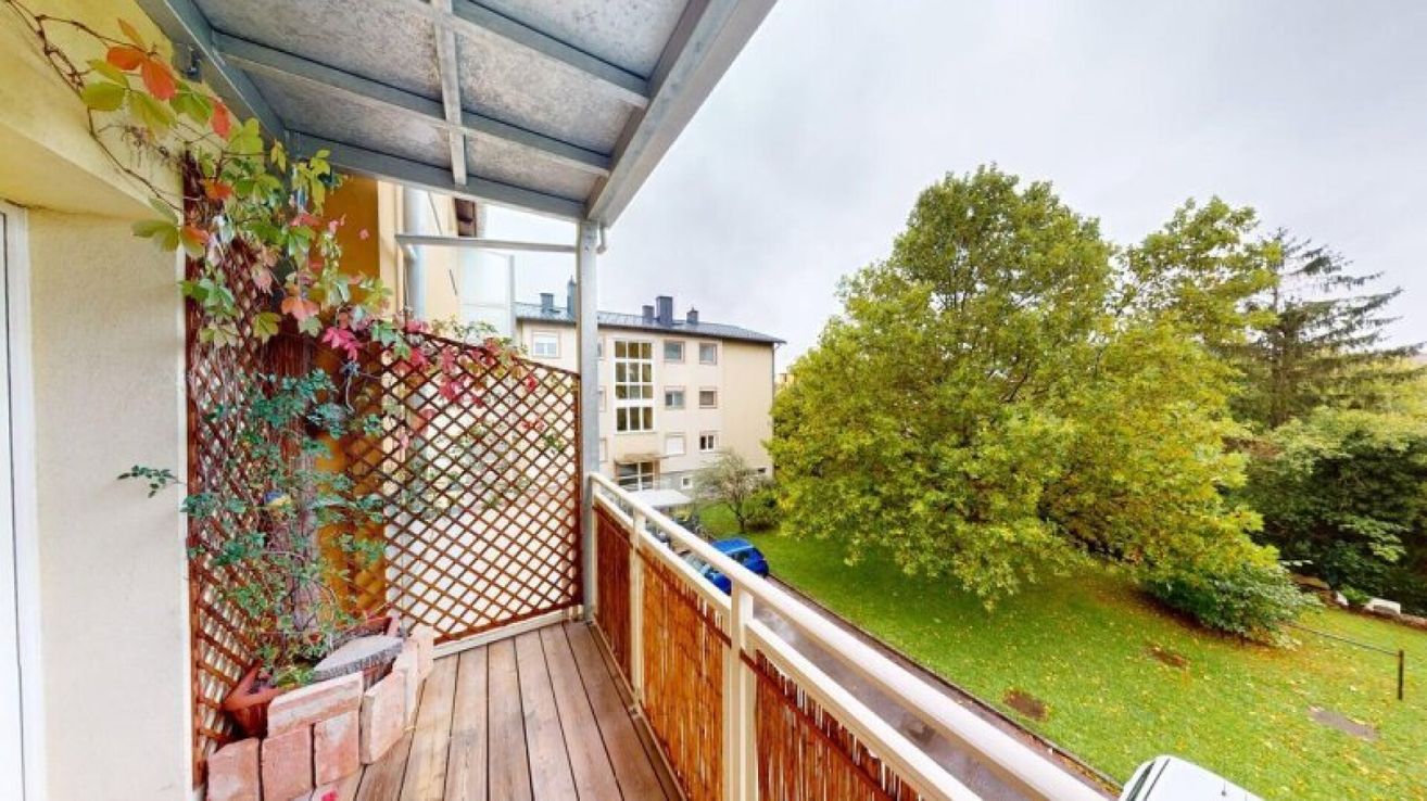 A balcony with a wooden railing and a metal gate, overlooking a grassy area with a tree and a blue car parked in front of a building.