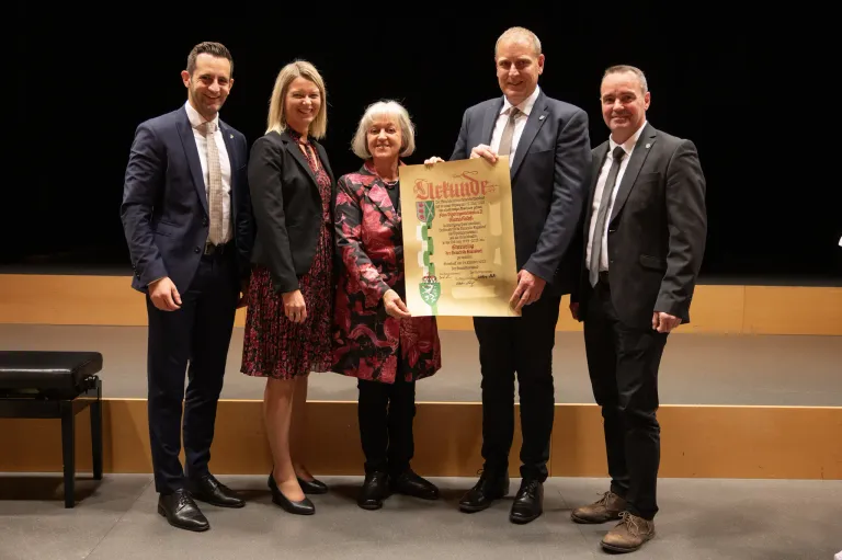 Fünf Personen in formeller Kleidung, zwei Frauen und drei Männer, stehen lächelnd auf einer Bühne. Eine Frau hält ein Zertifikat mit dem Text 'Stichtag' und einem Logo. Dahinter ist ein dunkler Hintergrund.