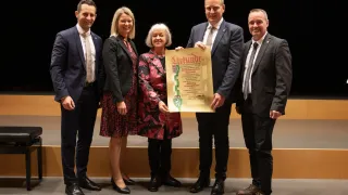 Fünf Personen in formeller Kleidung, zwei Frauen und drei Männer, stehen lächelnd auf einer Bühne. Eine Frau hält ein Zertifikat mit dem Text 'Stichtag' und einem Logo. Dahinter ist ein dunkler Hintergrund.