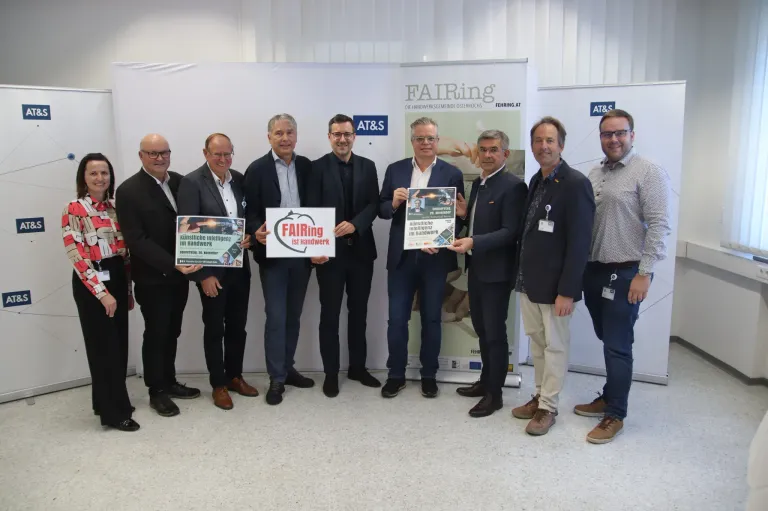 Eine Gruppe von Männern steht in einem Raum und hält Schilder. Ein Schild sagt FAIRing. Sie tragen alle formelle Kleidung. Dahinter ist ein Banner mit einem Logo und Text.