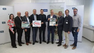 Eine Gruppe von Männern steht in einem Raum und hält Schilder. Ein Schild sagt FAIRing. Sie tragen alle formelle Kleidung. Dahinter ist ein Banner mit einem Logo und Text.