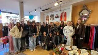 Eine Gruppe von Frauen und Männern posiert für ein Foto in einem Bekleidungsgeschäft. Einige stehen, andere sitzen. Der Laden hat eine gemütliche und festliche Atmosphäre mit Herbstdekorationen.