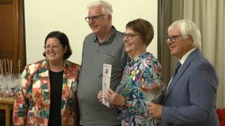 Vier lächelnde Erwachsene stehen zusammen. Eine Frau hält einen Blumenstrauß und ein Papier mit Schrift. Eine andere Frau hält ein Papier. Ein Mann hält einen Blumenstrauß. Sie alle tragen eine Brille.