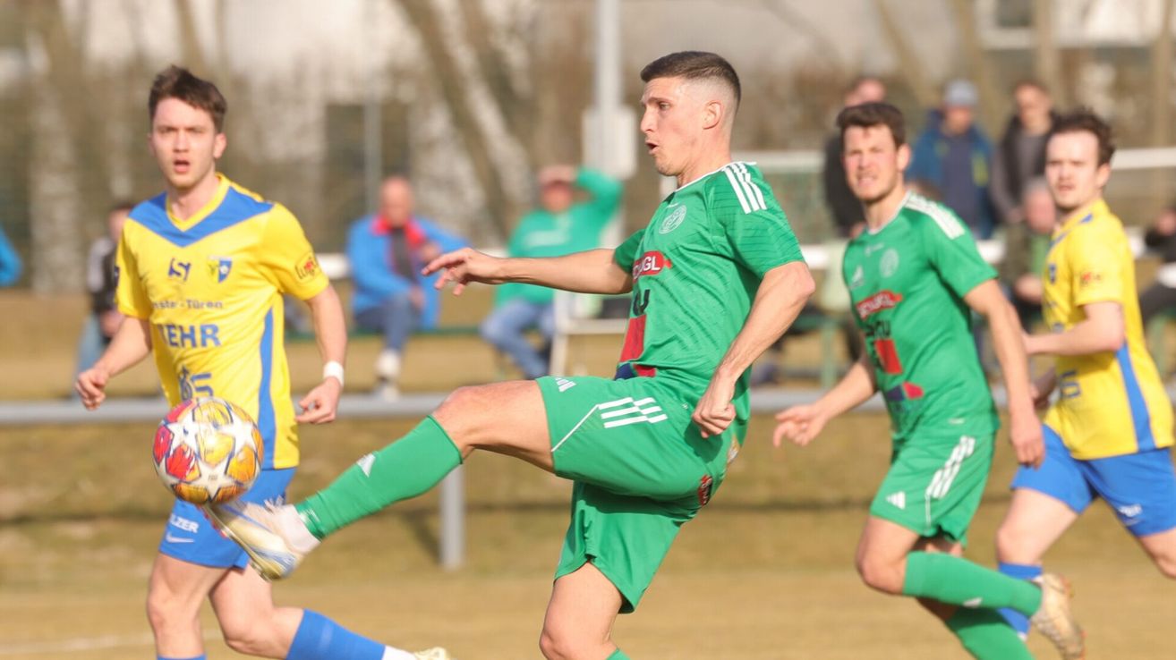 Ein Fußballspieler in einem grünen Trikot tritt den Ball auf einem Feld, während zwei andere Spieler und Zuschauer im Hintergrund zu sehen sind.