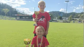 Zwei Kinder mit roten Shirts und Medaillen posieren für ein Foto mit Trophäen auf einem Rasenfeld. Hinter ihnen befindet sich ein Gebäude und ein hoher Lichtmast.