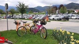 Drei Fahrräder mit Blumenkörben sind auf dem Rasen in der Nähe eines Parkplatzes mit vielen Autos geparkt, umgeben von Bäumen und Bergen.