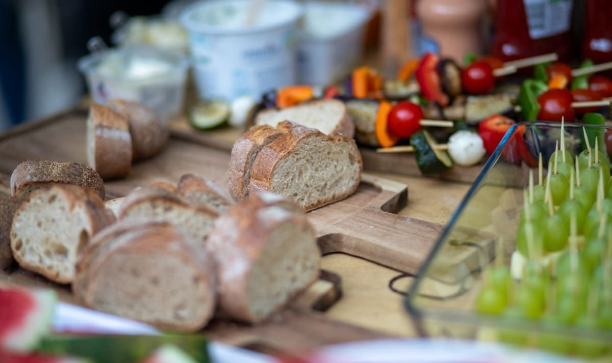 Ein Tisch ist mit geschnittenem Brot, einem Schneidebrett und Spießen mit Gemüse wie Tomaten, Paprika und Gurken bestückt. Im Hintergrund befinden sich Joghurtbecher und andere Gegenstände.